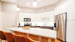 A modern kitchen with white cabinets, a stainless steel refrigerator, a microwave, and a large island with orange chairs.