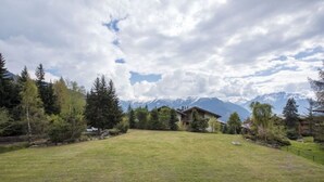 Un prato verdeggiante con un edificio al centro, circondato da alberi e montagne sullo sfondo.