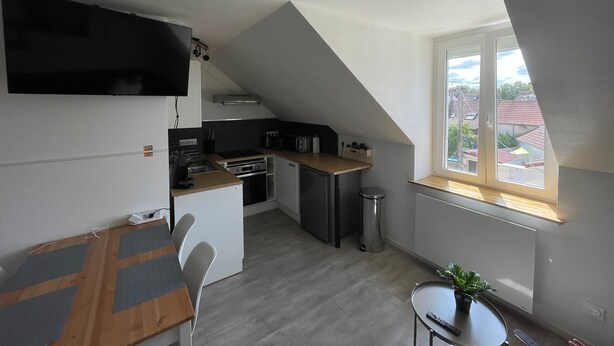 Et kompakt køkken med moderne hårde hvidevarer, et spiseområde med bord og stole, og et vindue med udsigt til bygninger.
