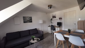 Et moderne, kompakt opholdsrum med en mørk sofa, et rundt bord med en plante, et skrivebord med en computer og et spiseområde med hvide stole.