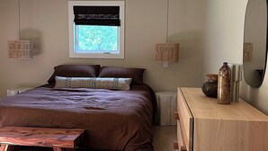 A modern bedroom with a large bed, bedside lamps, a wooden dresser, and a window with a view of greenery.