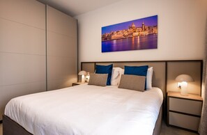A hotel room with a large bed, two bedside tables with lamps, a cityscape painting on the wall, and a view of the city from the window. - Charming 1BR Getaway, Great for Couples (Msida)