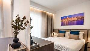 Une chambre d’hôtel avec un lit, une table de chevet, une petite table de nuit ornée d’un vase de fleurs séchées, et un tableau représentant un paysage urbain accroché au mur.