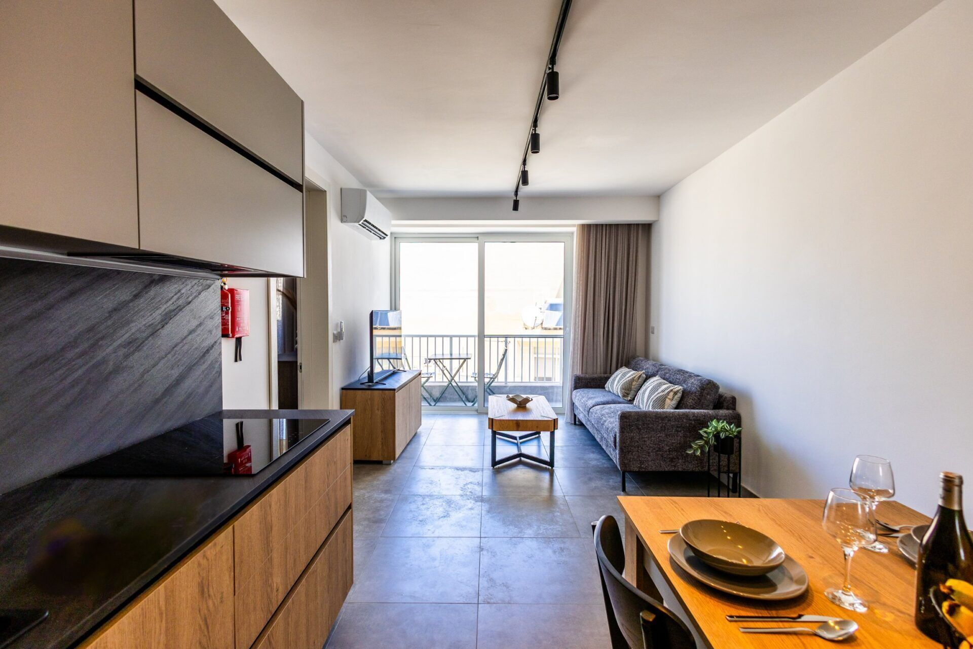 Eine moderne Küche mit schwarzer Arbeitsplatte und Holzschränken, ein Essbereich mit Holztisch und Stühlen, eine Sitzecke mit Sofa und ein Balkon mit Blick.