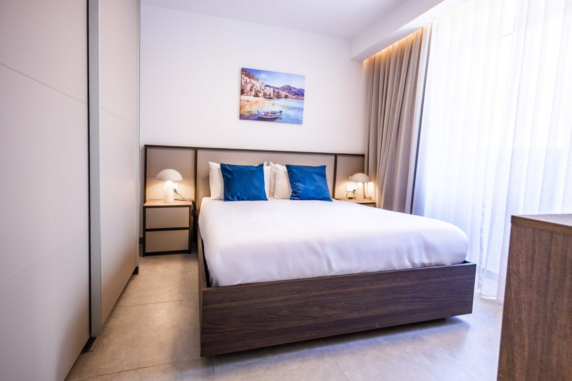 Quarto de hotel moderno com uma cama grande, duas mesas de cabeceira com luminárias, um criado-mudo e um quadro na parede.