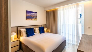 A hotel room with a large bed, a nightstand with a lamp, a painting on the wall, and a view of the outdoors through sheer curtains.