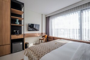 A modern hotel room with a large bed, a desk with a computer, a small fridge, and a TV mounted on the wall.