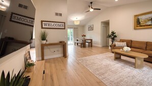A spacious living room with a wooden floor, a sofa, a coffee table, and a dining area with a bench and table. There are two welcome signs on the wall, a ceiling fan, and a painting on the wall.