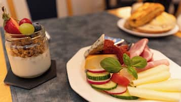 A breakfast setting with a plate of sliced fruits, cheese, and ham, and a jar of yogurt with granola and fruit on a table.