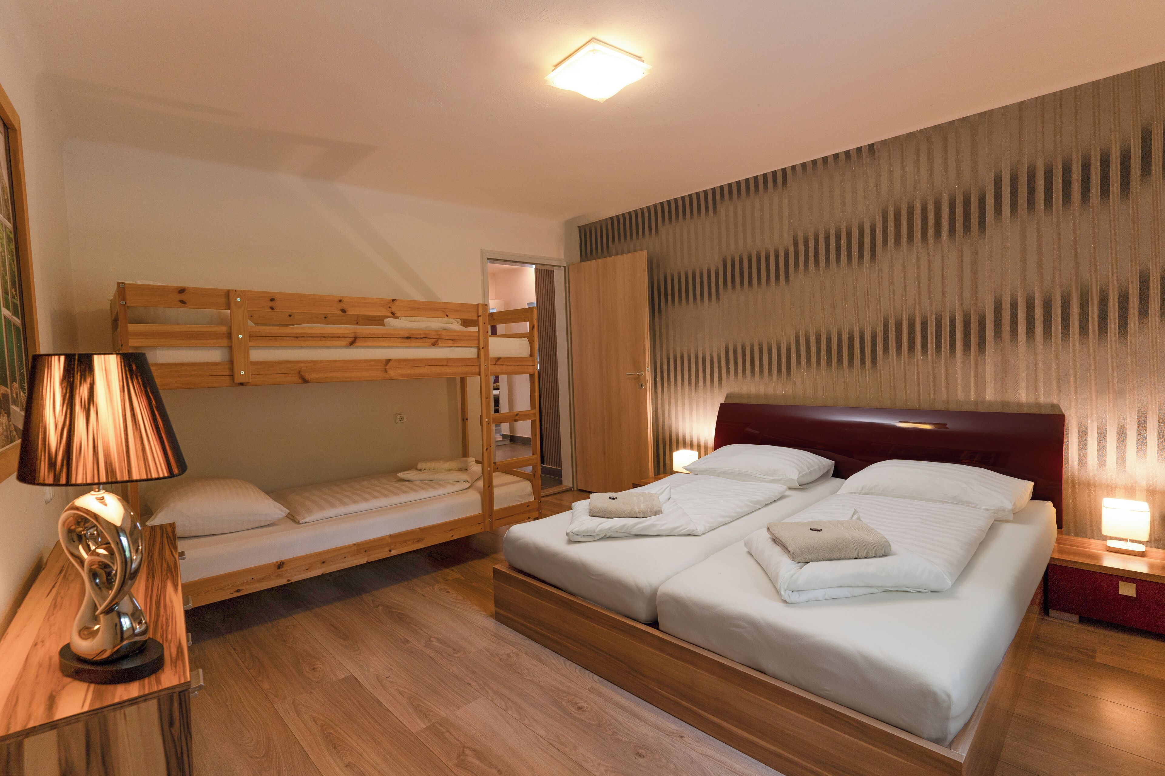 A room with a bunk bed, a single bed, a bedside table with a lamp, and a wooden floor.
