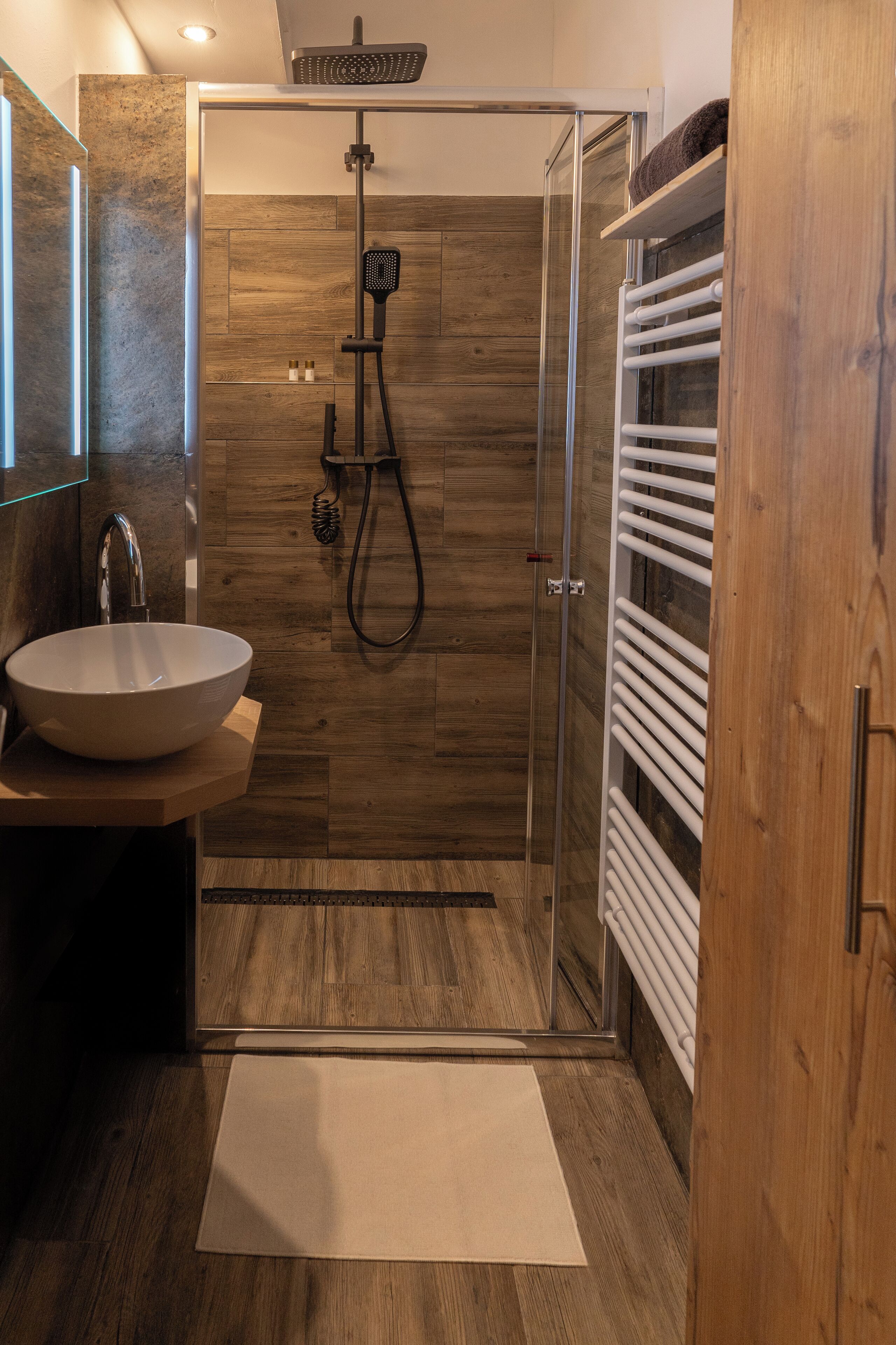 Un baño moderno con cabina de ducha de vidrio, suelo de madera, lavamanos blanco y un perchero.