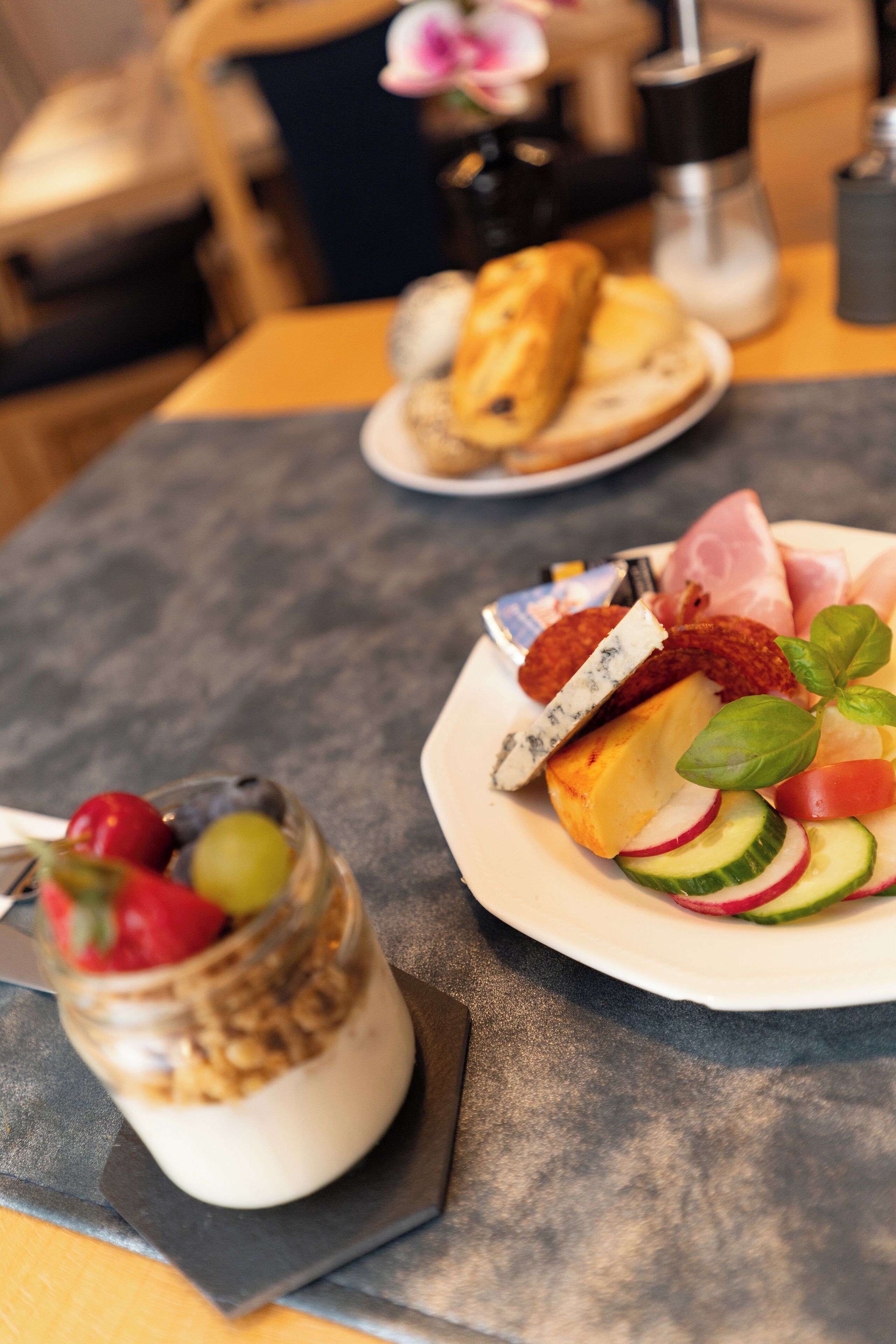 Una mesa con diversos alimentos, incluyendo un vaso de yogur con frutas, un plato de queso, carnes y verduras, y una baguette.