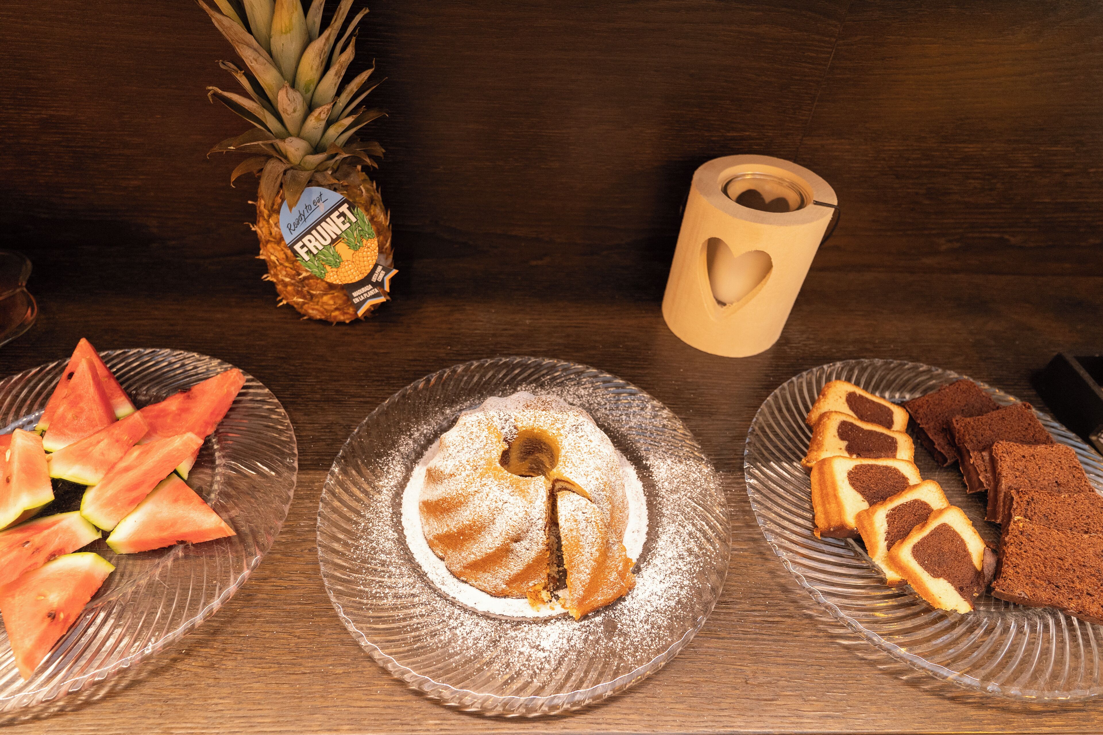 Ein Holztisch mit einer Glasplatte, auf der eine Ananas, Wassermelonenscheiben und eine Kuchenplatte mit Keksen liegen.