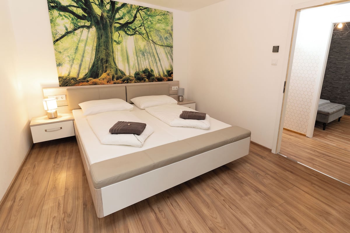 Quarto moderno com uma cama grande, mesa de cabeceira com abajur e um mural de floresta na parede.