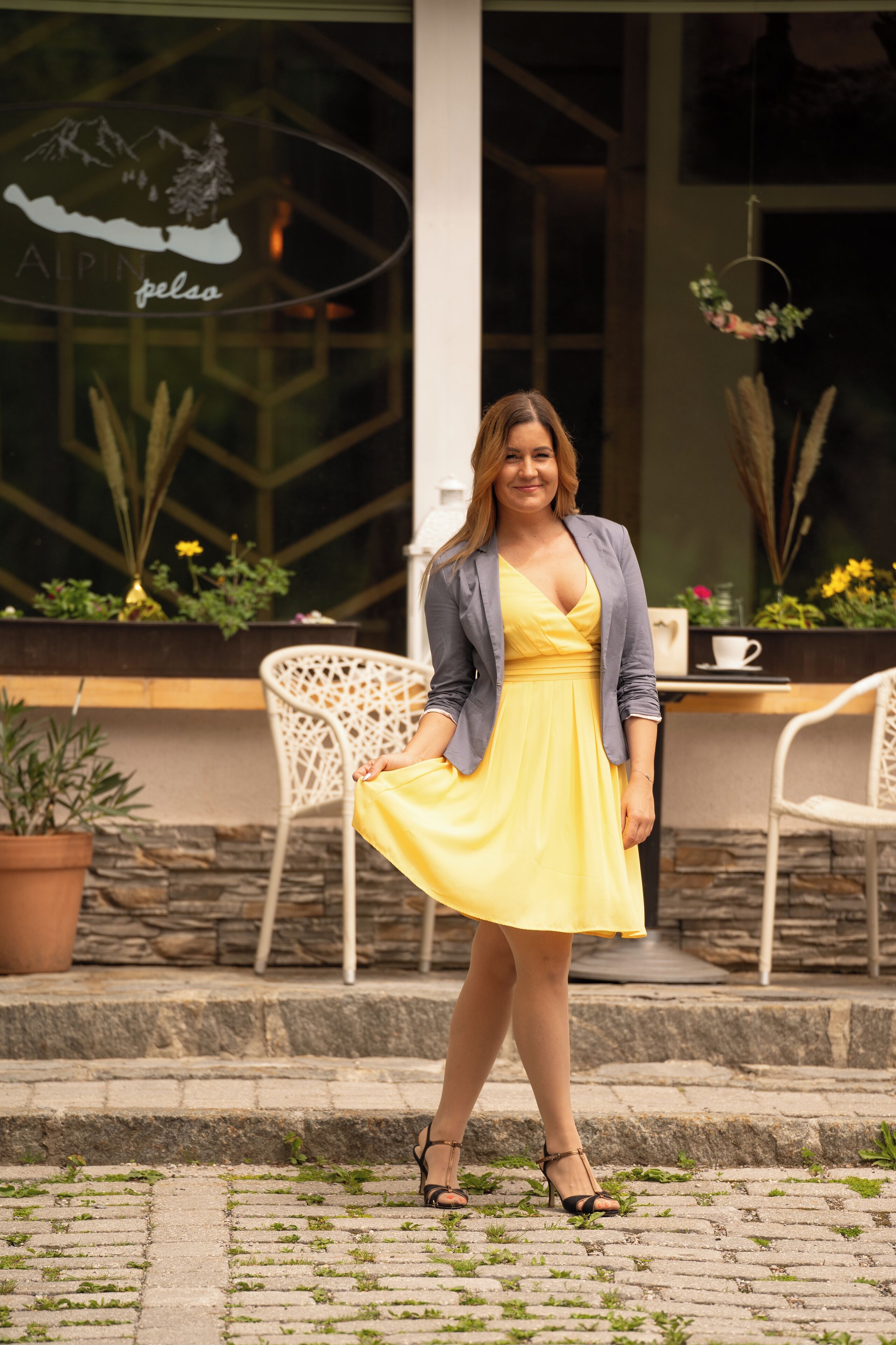 A woman in a yellow dress standing on a cobblestone path with a chair and table in the background.