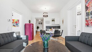 Un salon moderne avec deux canapés gris, une table basse ronde en bois foncé et un vase éclatant rempli de fleurs violettes. Un réfrigérateur rose de style rétro est visible au fond.