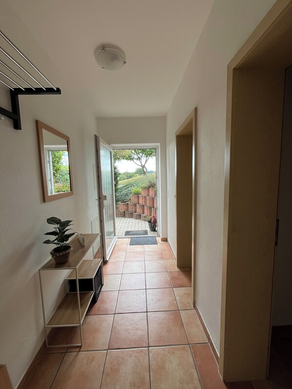 A hallway with a tiled floor, a potted plant, a mirror, and a door leading to an outdoor area with a brick wall and greenery.