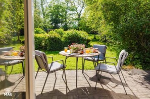 Een terras buiten met een ronde tafel en vier stoelen, omgeven door weelderige begroeiing en een terrasvloer van baksteen.