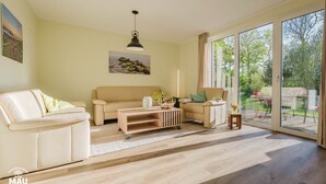 A spacious living room with a beige sofa, a wooden coffee table, and a large sliding glass door leading to a garden. - Holiday Farm Mau - House 1 (Fehmarn)