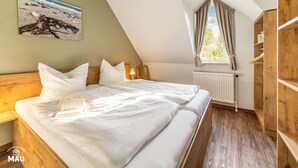 A bedroom with a bed, a nightstand, a wooden wardrobe, a window with curtains, and a painting on the wall. - Holiday Farm Mau - House 1 (Fehmarn)