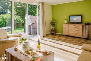 A modern living room with a large sliding glass door leading to a garden, a wooden cabinet with a TV, and a cozy seating area with a coffee table. - Holiday Farm Mau - House 1 (Fehmarn)