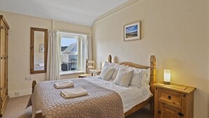 Een slaapkamer met een groot bed, een nachtkastje met een lamp, een houten garderobe, een raam met gordijnen en een lijst met een foto aan de muur.