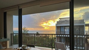 Een balkon met uitzicht op de ondergaande zon boven de oceaan, een eethoek met een tafel en stoelen, en een modern gebouw op de achtergrond.