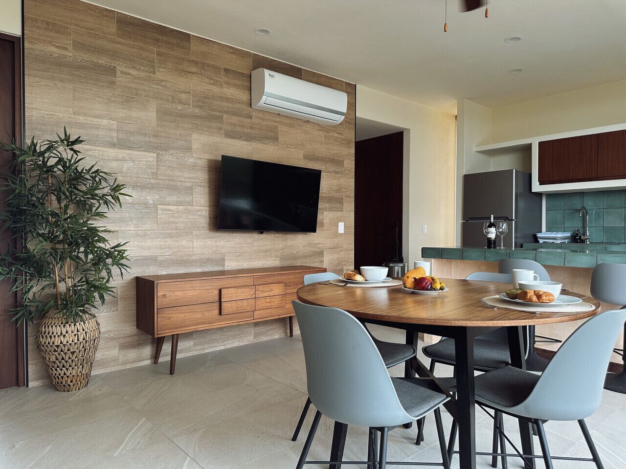 A modern dining area with a wooden table, grey chairs, a flat-screen TV mounted on the wall, and a kitchen in the background.