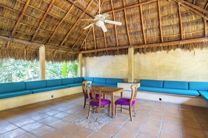 A room with a thatched roof, blue cushioned seating, a wooden table with purple chairs, and a tiled floor.