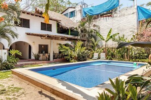 Una piscina con borde de azulejos, rodeada por un jardín con diversas plantas y un edificio con techo de paja.