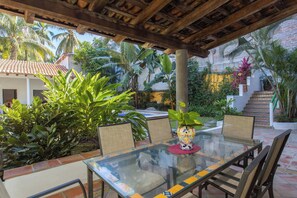 Un patio exterior con una mesa y sillas de vidrio, rodeado de abundante follaje y un muro de ladrillo.