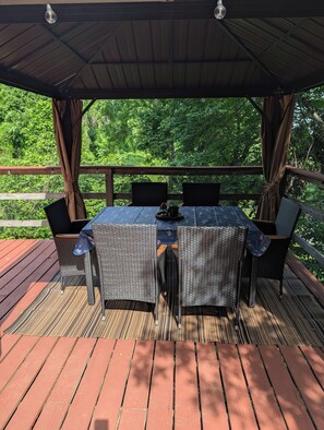 Una terraza techada al aire libre con una mesa y sillas, rodeada por una cerca de malla, con vista a una zona verde exuberante.