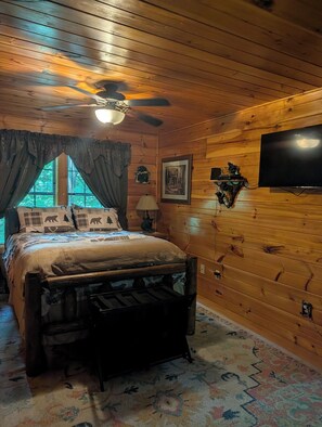 Une chambre dans un chalet en bois, avec un lit, un ventilateur de plafond, une télévision et une fenêtre avec des rideaux.