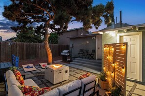 Un patio trasero con terraza, mobiliario exterior, una parrilla y una valla de madera.