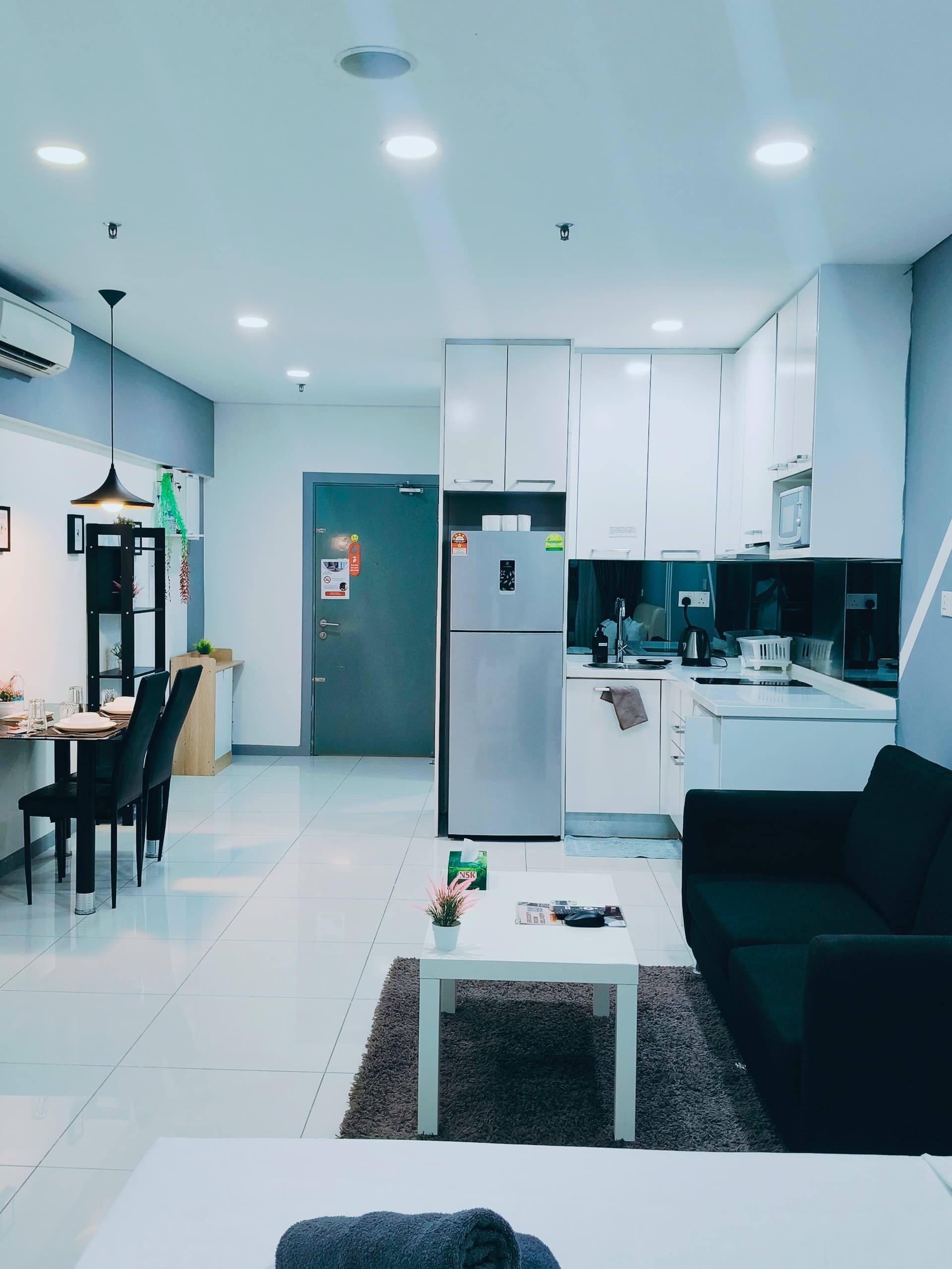 A modern kitchen with white cabinets, a built-in oven, and a refrigerator. There is a dining area with a table and chairs, and a living room with a sofa and a coffee table.