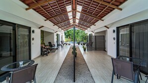 A covered outdoor area with a central pathway, seating arrangements, and a view of greenery in the background.