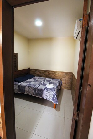 Una habitación pequeña con cama individual, cabecera de madera, colcha estampada y aire acondicionado de pared.