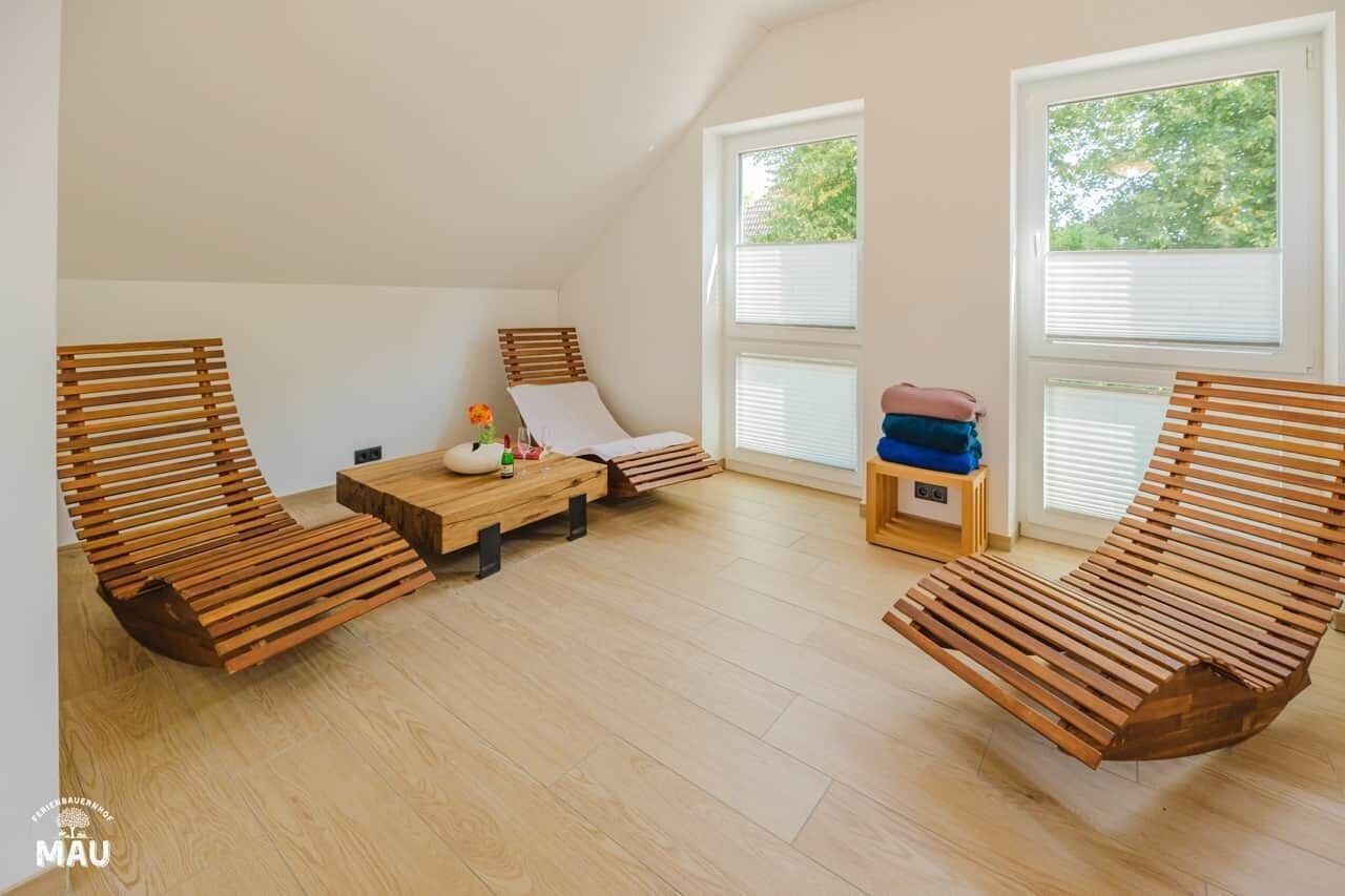 Una habitación con dos sillones de madera, una mesita y un banco. Hay dos ventanas con persianas, y un taburete con un bolso.