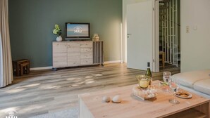 Uma sala de estar moderna com piso de madeira, uma TV em um móvel de madeira, uma mesa de jantar com comida e bebidas, e uma porta deslizante que dá para um armário.