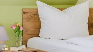 Cama bem arrumada com roupa de cama e almofadas brancas, cabeceira de madeira, mesa de cabeceira com abajur e um livro, e um vaso com tulipas.