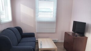 A living room with a dark blue sofa, a wooden coffee table, a TV on a wooden cabinet, and two windows with blinds. - Sarah Cottage - 226 Cottage Road, Steeprock , Manitoba (Grahamdale)