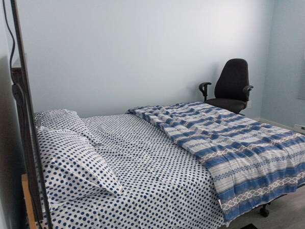 Una habitación pequeña con cama individual, colcha de lunares y cuadros, una silla de oficina negra y paredes azul claro.