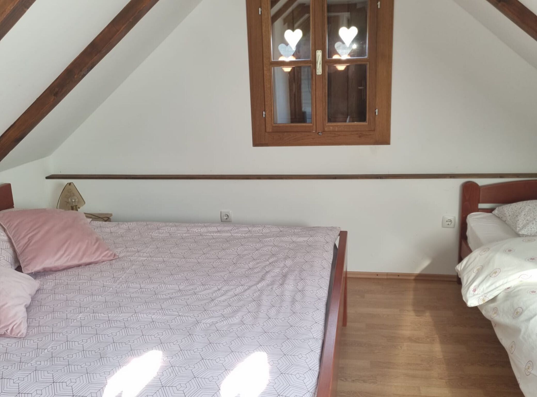 A bedroom with a sloped ceiling, a wooden bed frame, a window with heart-shaped decorations, and a bedside lamp.