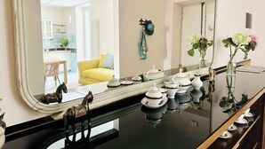 A room with a large mirror reflecting a dining area, a black table with white and gold tea sets, and a wall-mounted hat rack with a green hat. - TrasteverHome -Cisterna Secrets at the Heart of Trastevere (Rome)