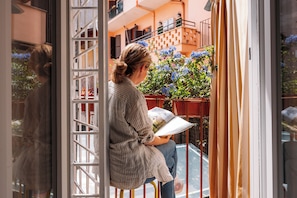 Une personne assise sur une chaise près d’une fenêtre, avec vue sur un balcon orné de plantes.