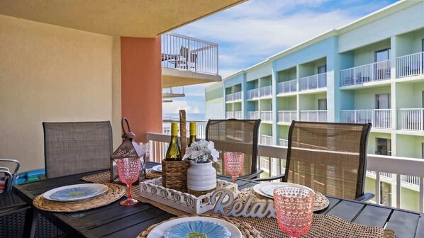 A balcony with a table set for dining, featuring wicker chairs, a tray with plates, glasses, and bottles. The background includes a multi-story building with balconies. - Hot Tub•Oceanfront •Heated Pools•King•Cable TV•W/D (Orange Beach)