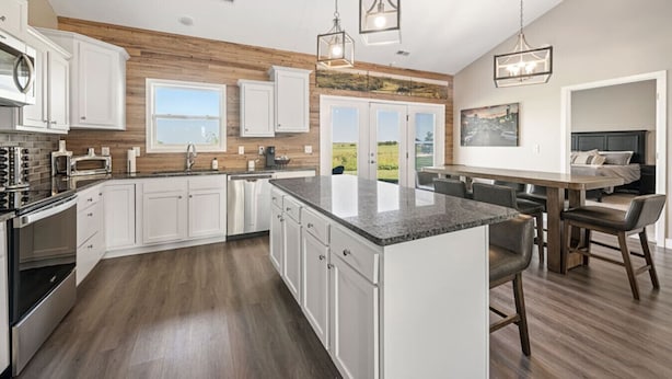 A modern kitchen with white cabinets, a granite countertop, and a large island. There are hanging lights and a view of the outdoors through the windows.