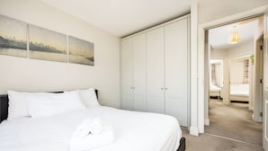 Una cama bien tendida con sábanas blancas y una toalla enrollada, un armario blanco grande y una pintura de paisaje urbano en la pared.