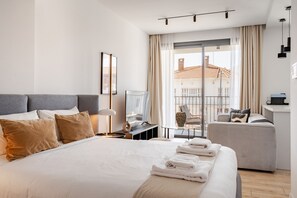 A modern hotel room with a large bed, a desk with a chair, a sofa, and a balcony with a view of buildings. - Yamas Urban Living Olympia Suites (Limassol)
