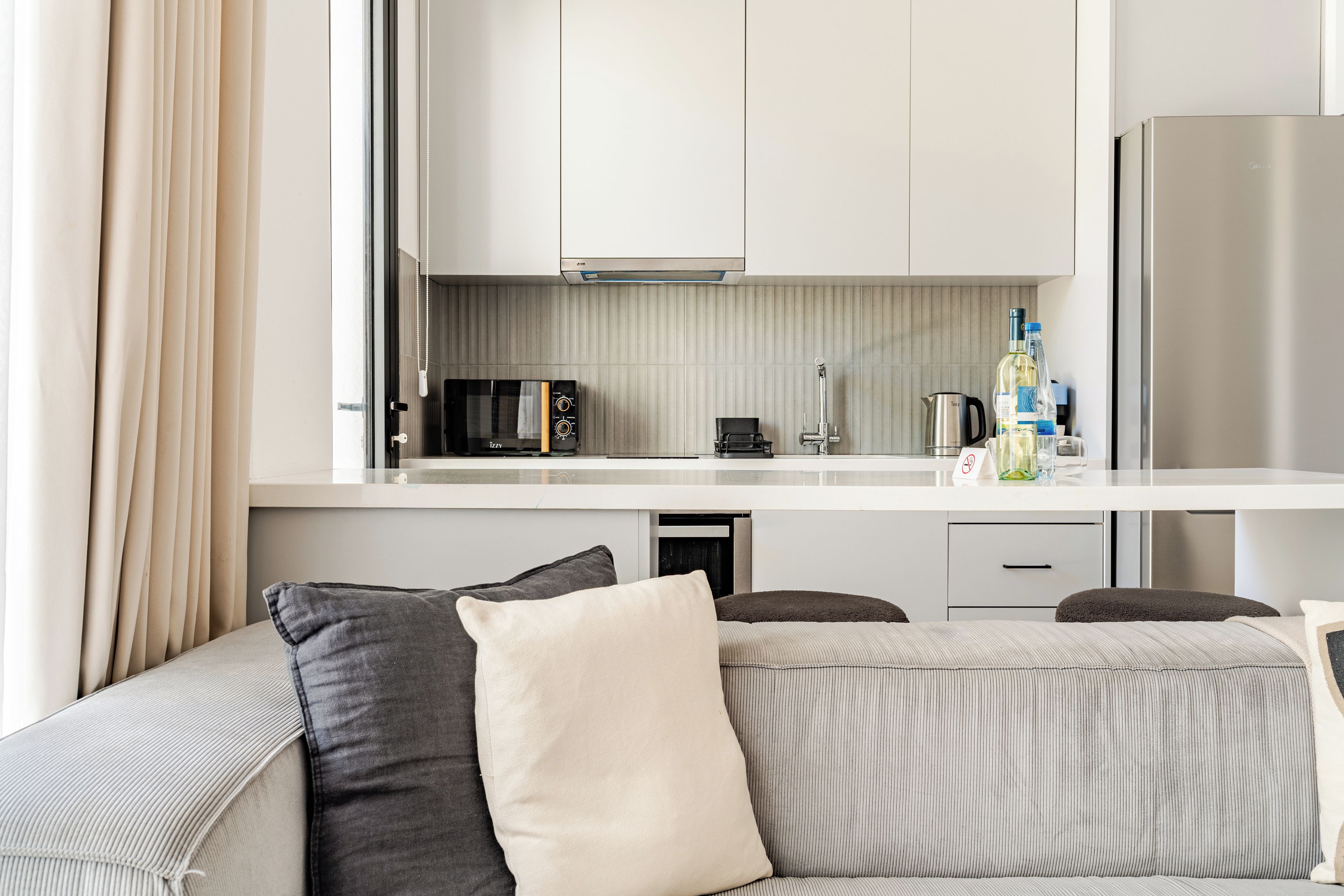 Een moderne kitchenette met een magnetron, waterkoker en koffiezetapparaat. Er is een bank met kussens en een glazen salontafel.